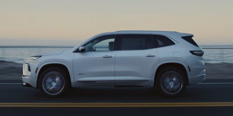 A white Buick Envista SUV, shown in profile, driving on a coastal road next to the ocean at dusk