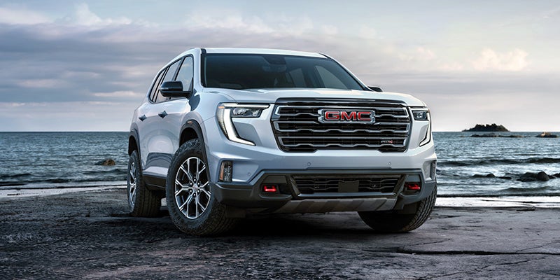 A white GMC SUV parked on a dark, rocky beach, with the ocean and a cloudy sky in the background