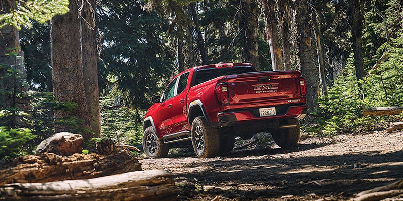 Rear view of a red GMC Canyon pickup truck driving away on a dirt road through a sunny forest