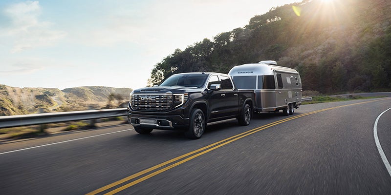 Black GMC Sierra pickup truck towing a silver Airstream trailer on a winding road with mountains and forests at sunset