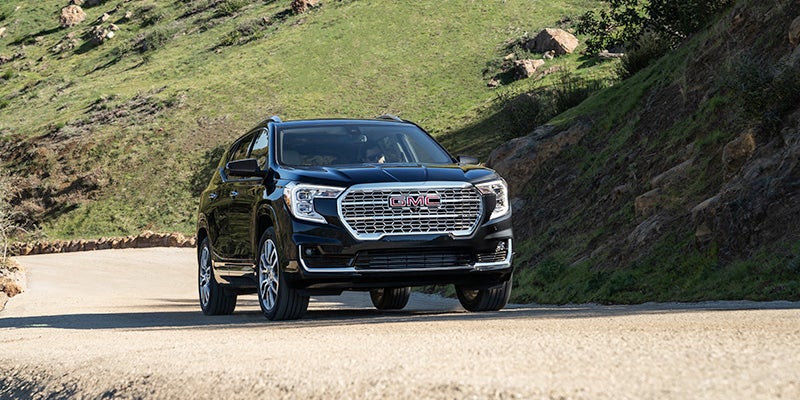 A black GMC Acadia SUV driving on a winding dirt road through a green, rocky hillside