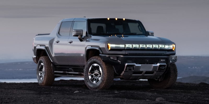 Front view of a dark gray GMC Hummer EV parked on rough terrain against a cloudy, light gray sky