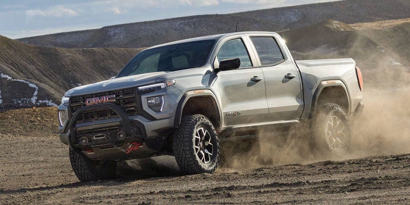 A light gray GMC Canyon AT4X pickup truck driving on a dirt road, kicking up dust behind its rear tires
