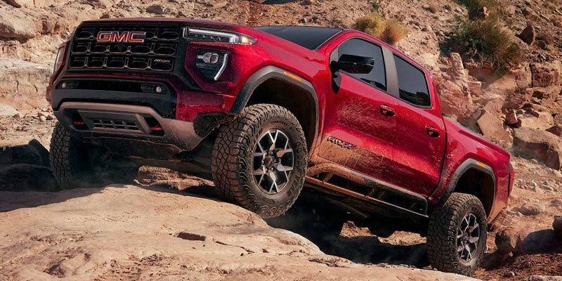 A red GMC pickup truck with large off-road tires angles up a rocky, dirt trail, showing its front left