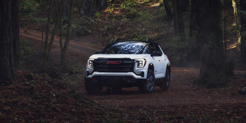 A white GMC SUV with black accents drives on a dirt road through a dark forest.